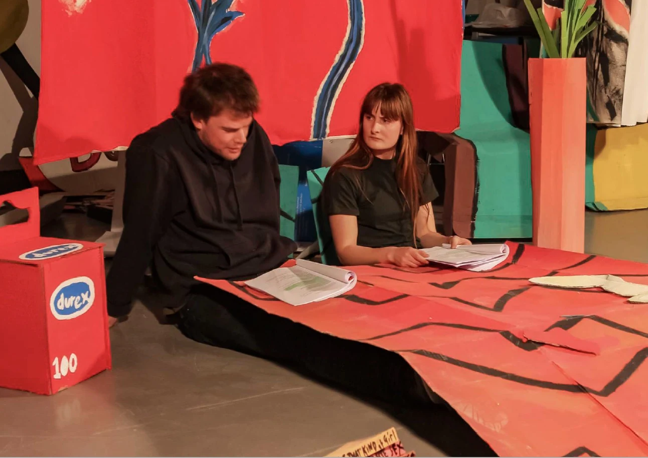 Two performers reading scripts beside red cardboard bedding on stage.
