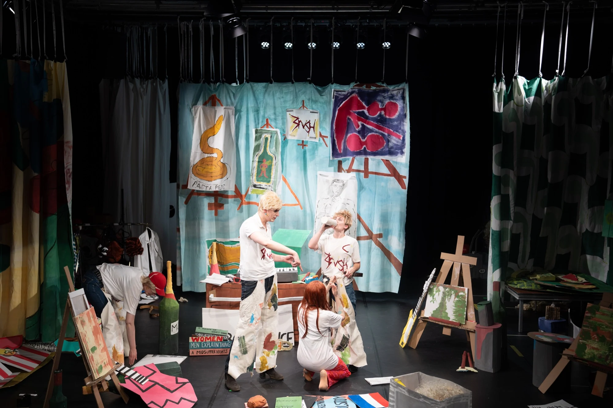 Performers acting among handmade paintings, easels and cardboard props.