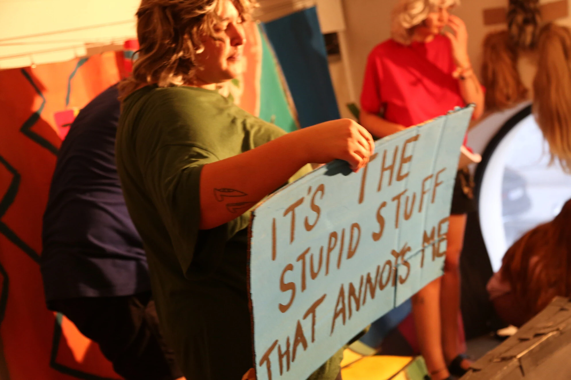 A performer in green holding a blue sign reading It's the stupid stuff that annoys me.