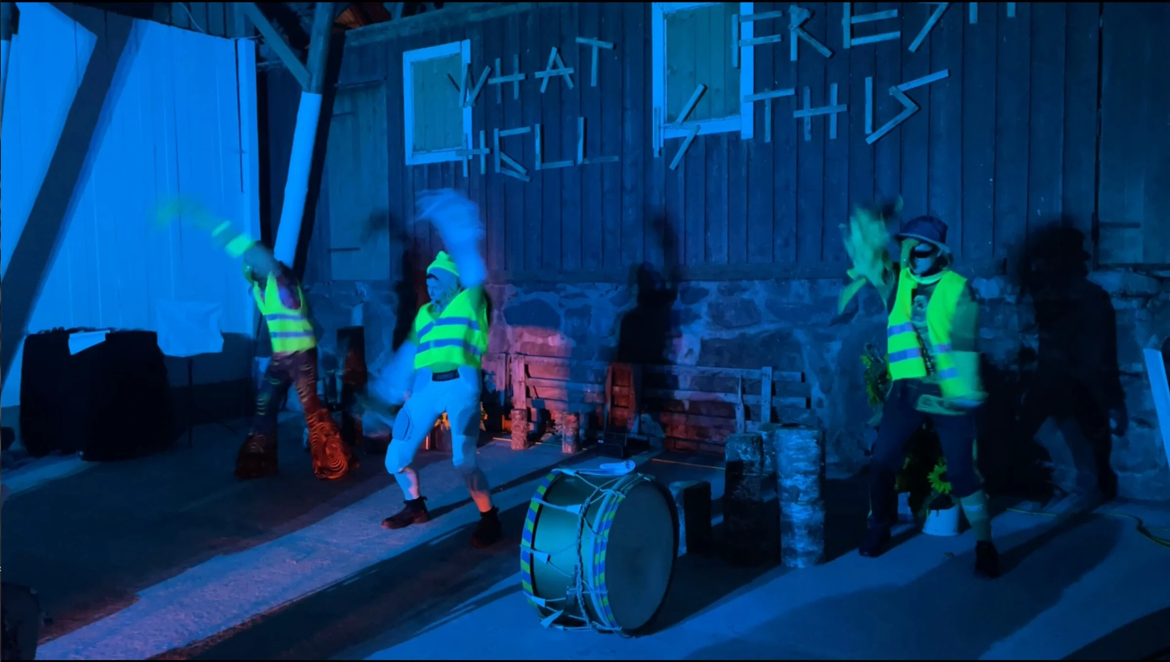 Three performers in fluorescent vests raising their arms beside a drum.