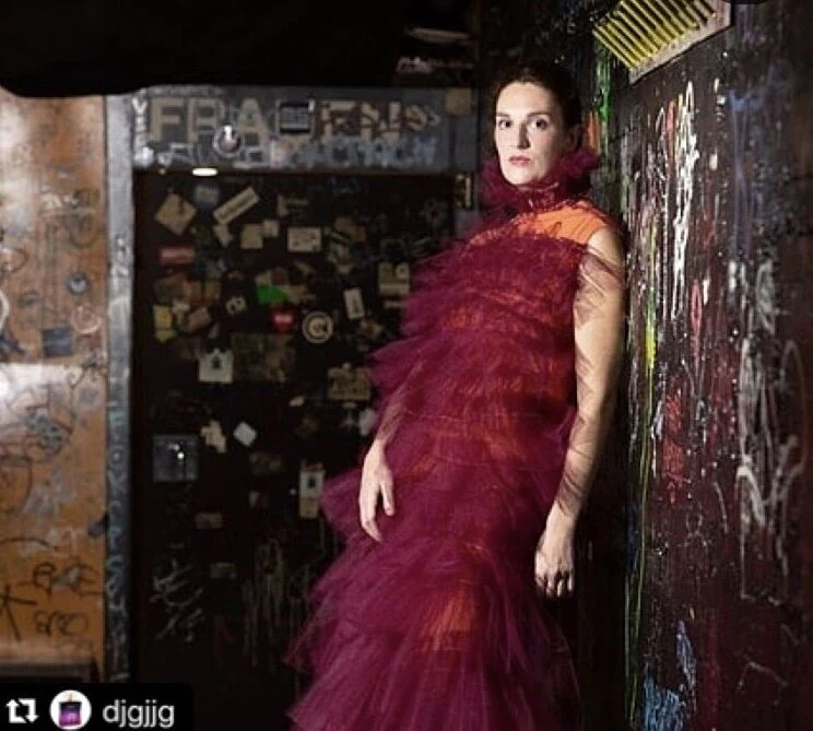 A performer in a burgundy tulle costume posing against a graffiti-covered wall.