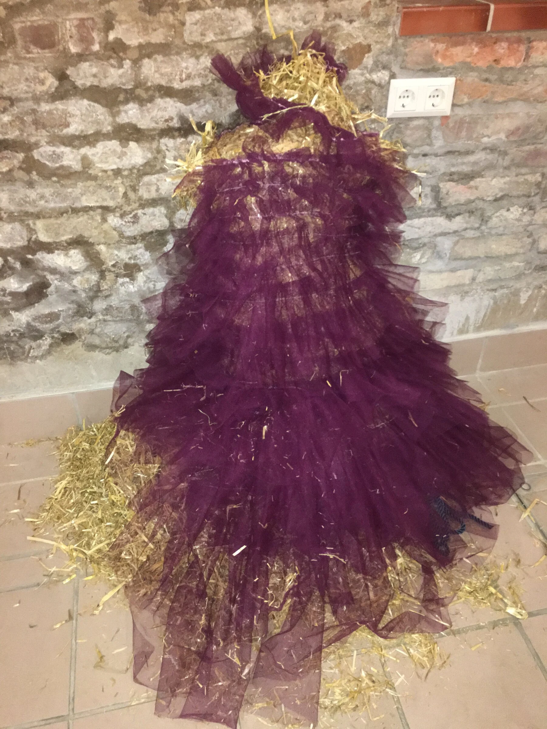 A burgundy tulle costume filled with straw beside a brick wall.