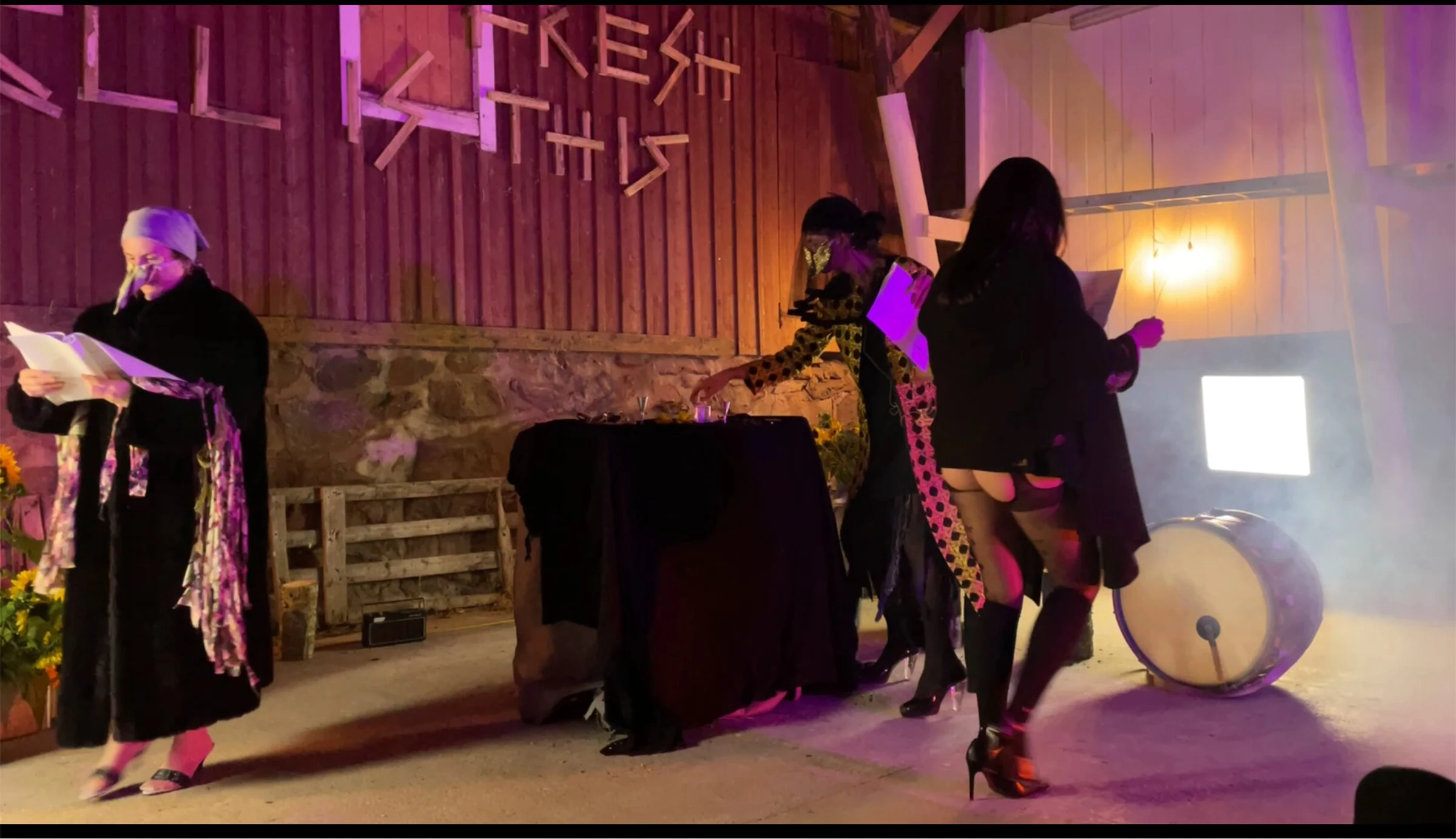 Masked performers reading scripts beside a black-draped table and a drum.