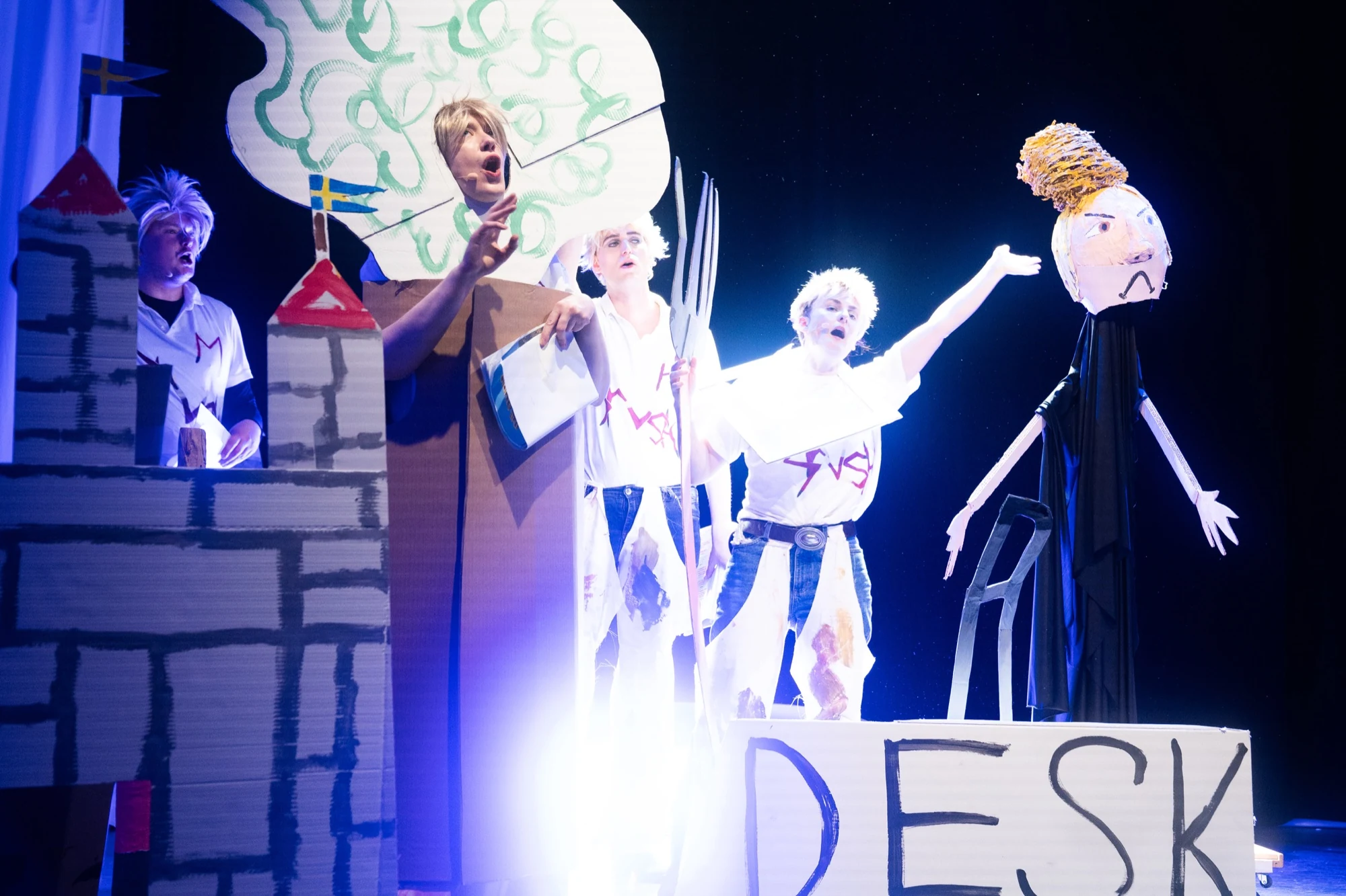 Performers singing beside a cardboard castle, tree and puppet under bright stage lighting.