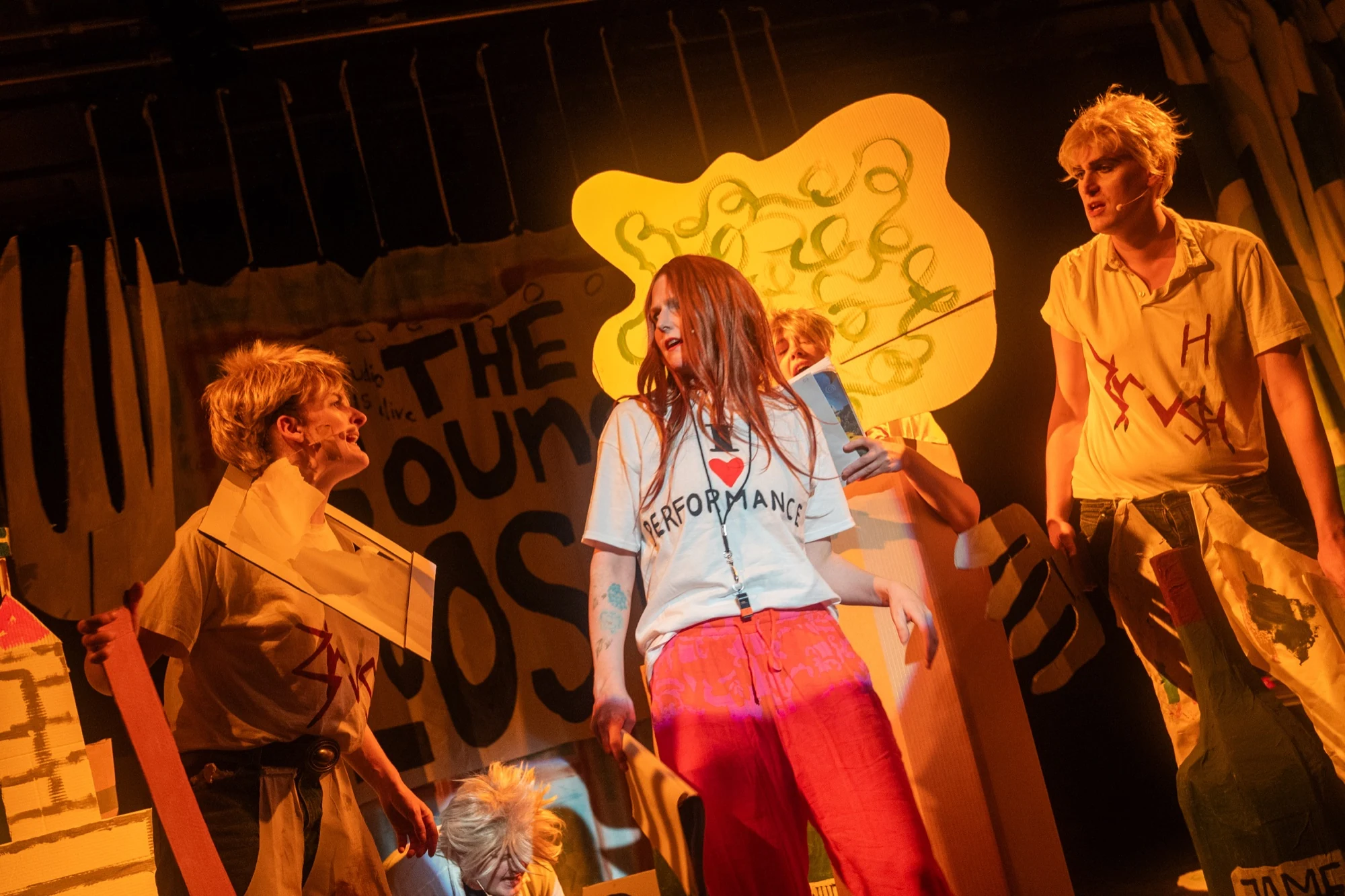 Charlotte surrounded by performers and cardboard scenery under warm stage lighting.