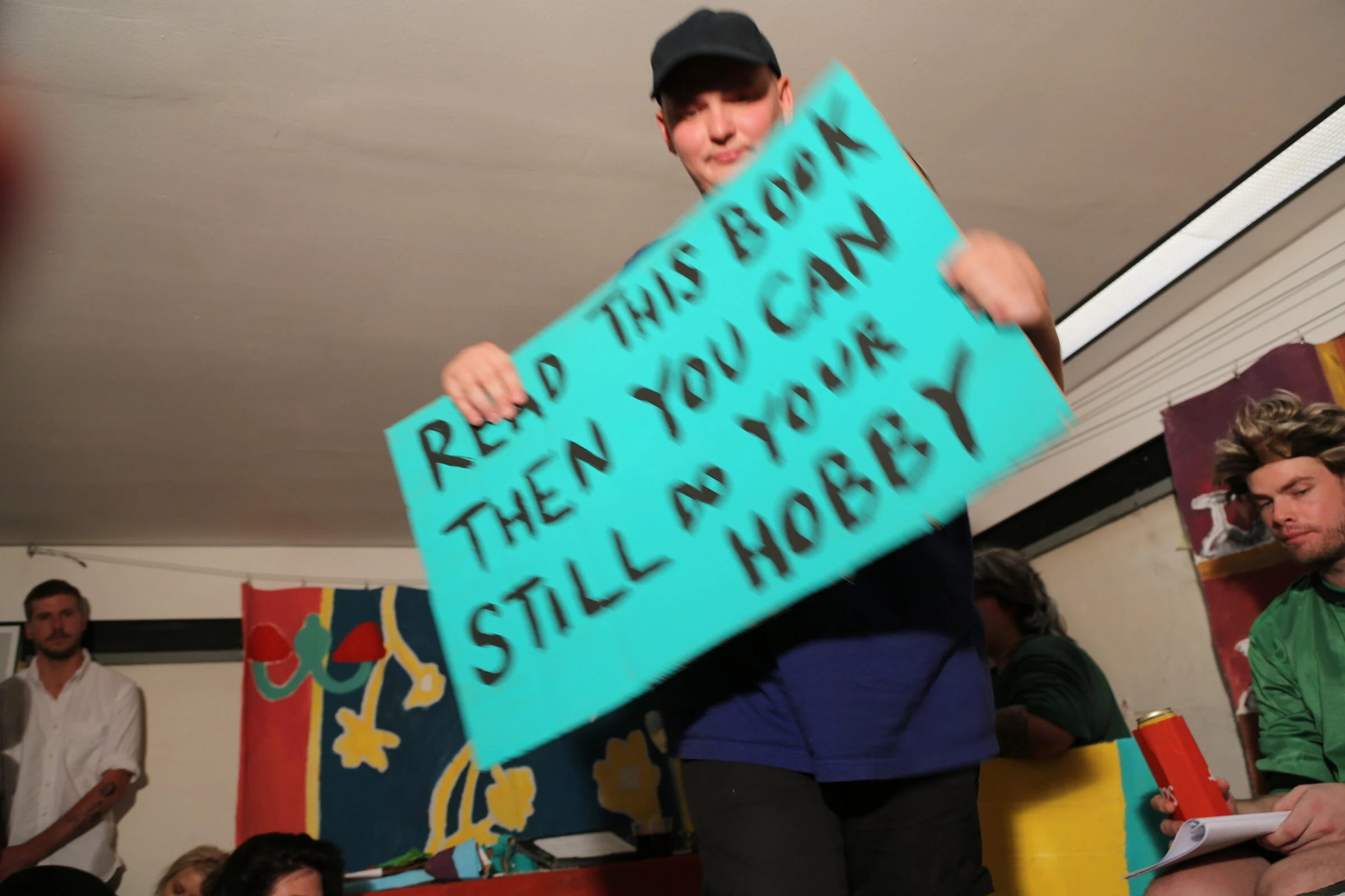A performer holding a turquoise cardboard sign reading Read This Book Then You Can Still Do Your Hobby.