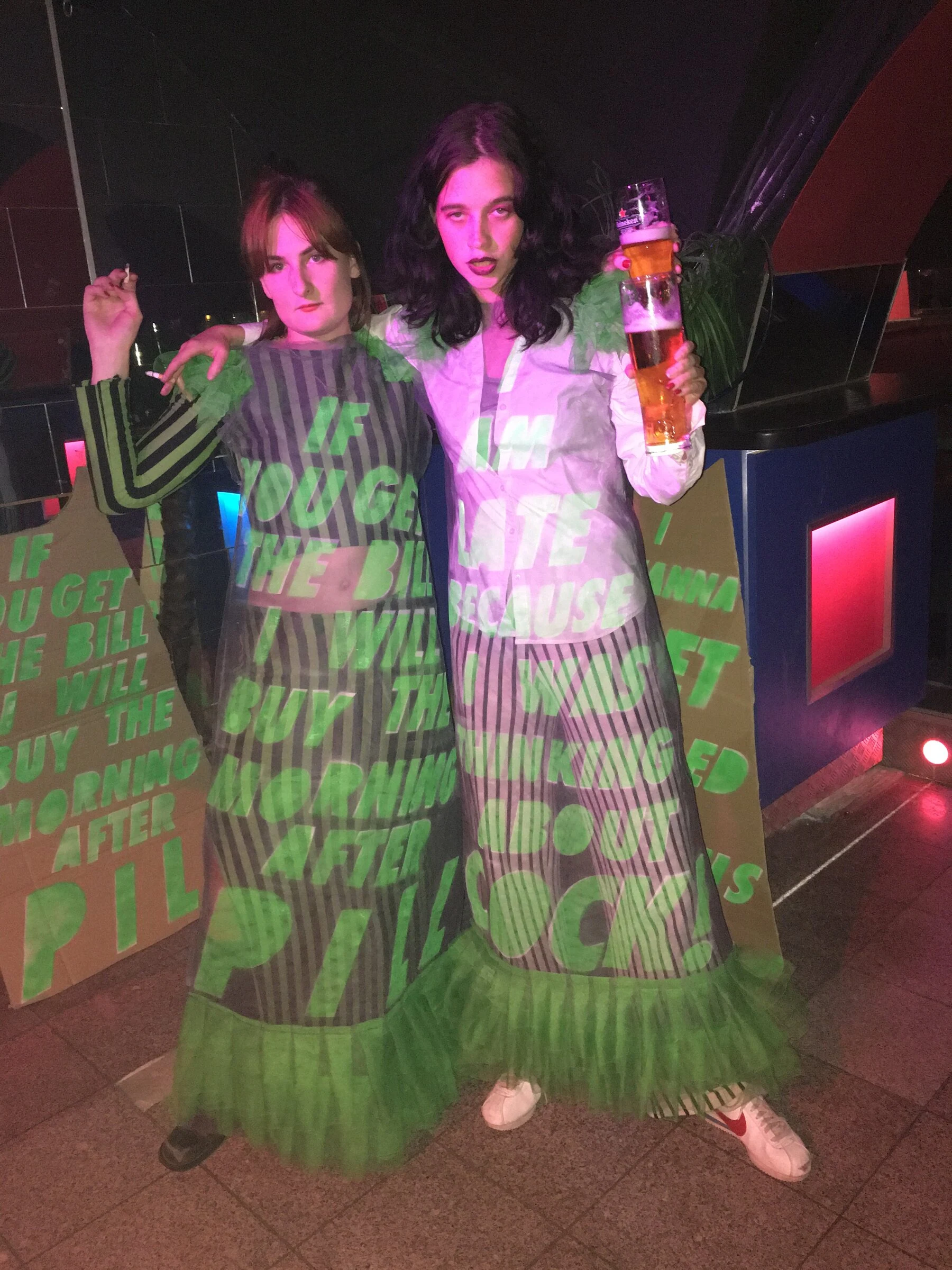Two performers wearing dresses with projected green lettering and holding stacked beer glasses.