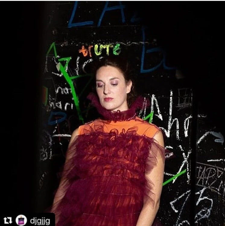 A close-up of a performer in a burgundy tulle costume against a graffiti-covered wall.