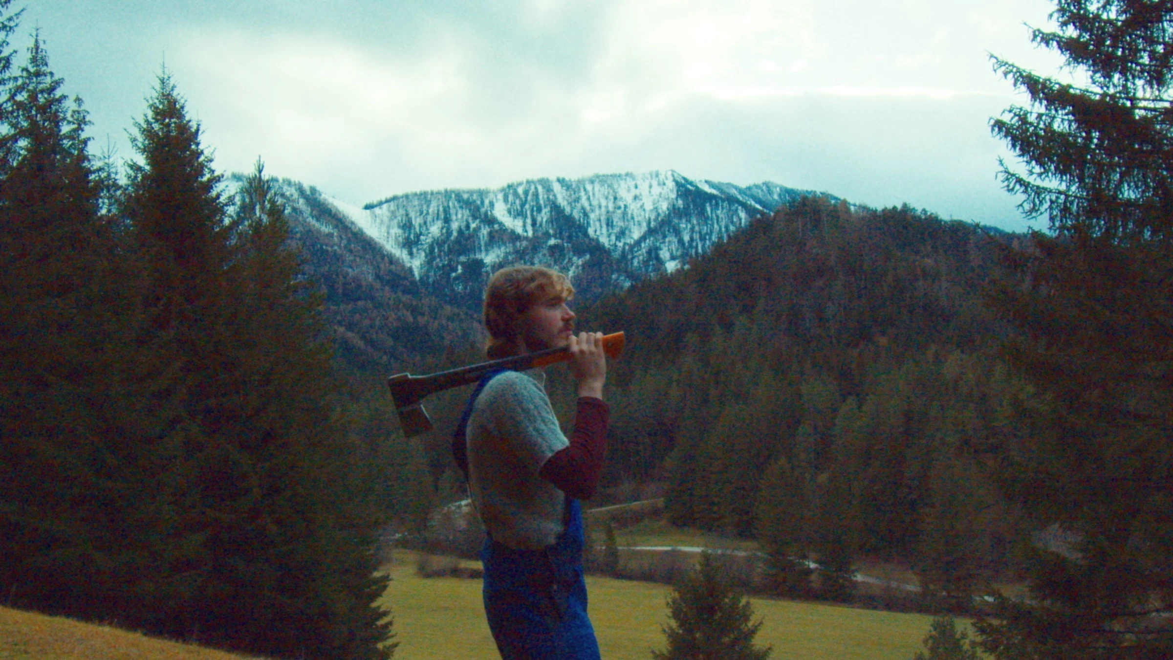 A performer in blue overalls carrying an axe against a mountain landscape.
