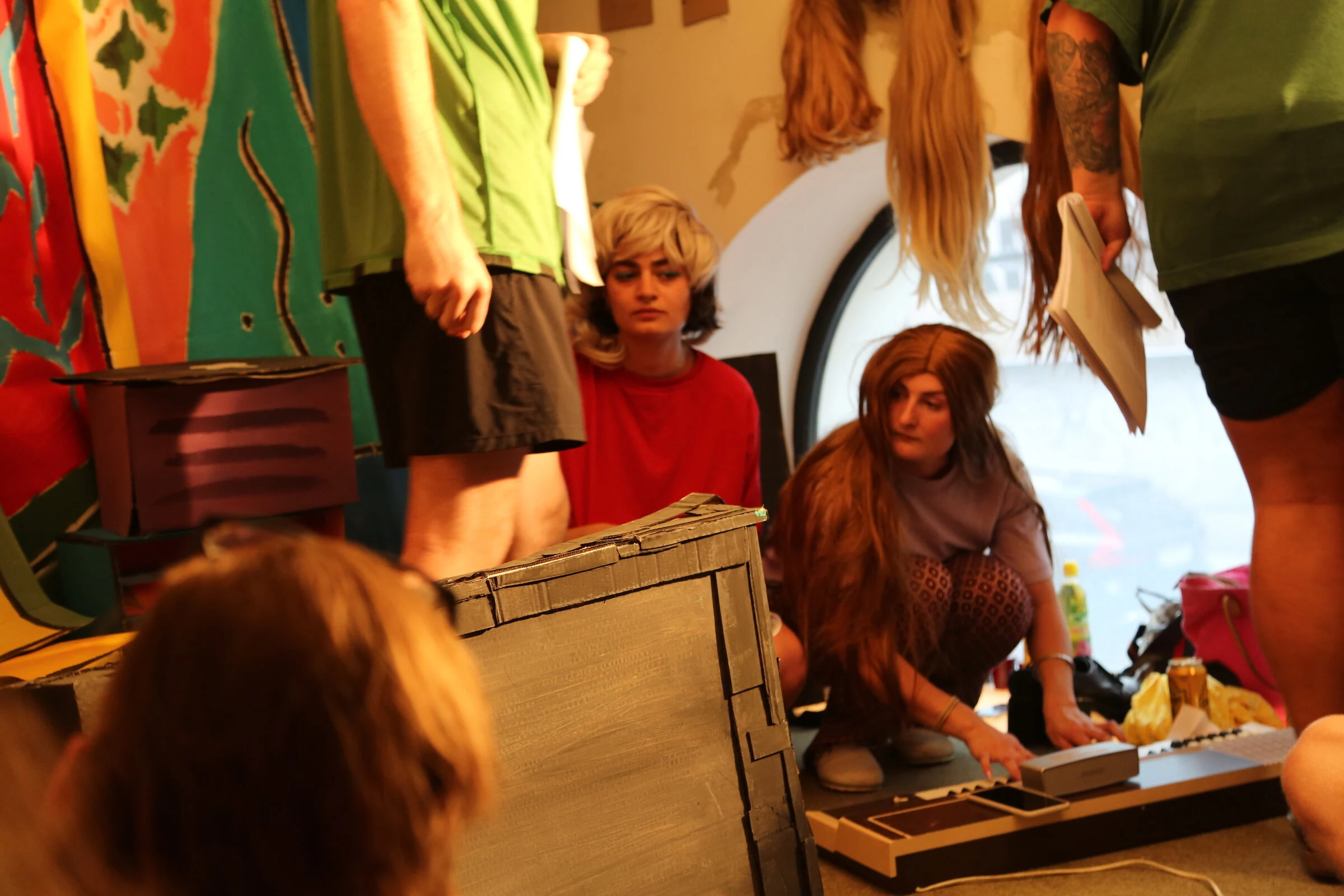 Two performers in wigs crouching beside a keyboard and cardboard television while other performers stand nearby.