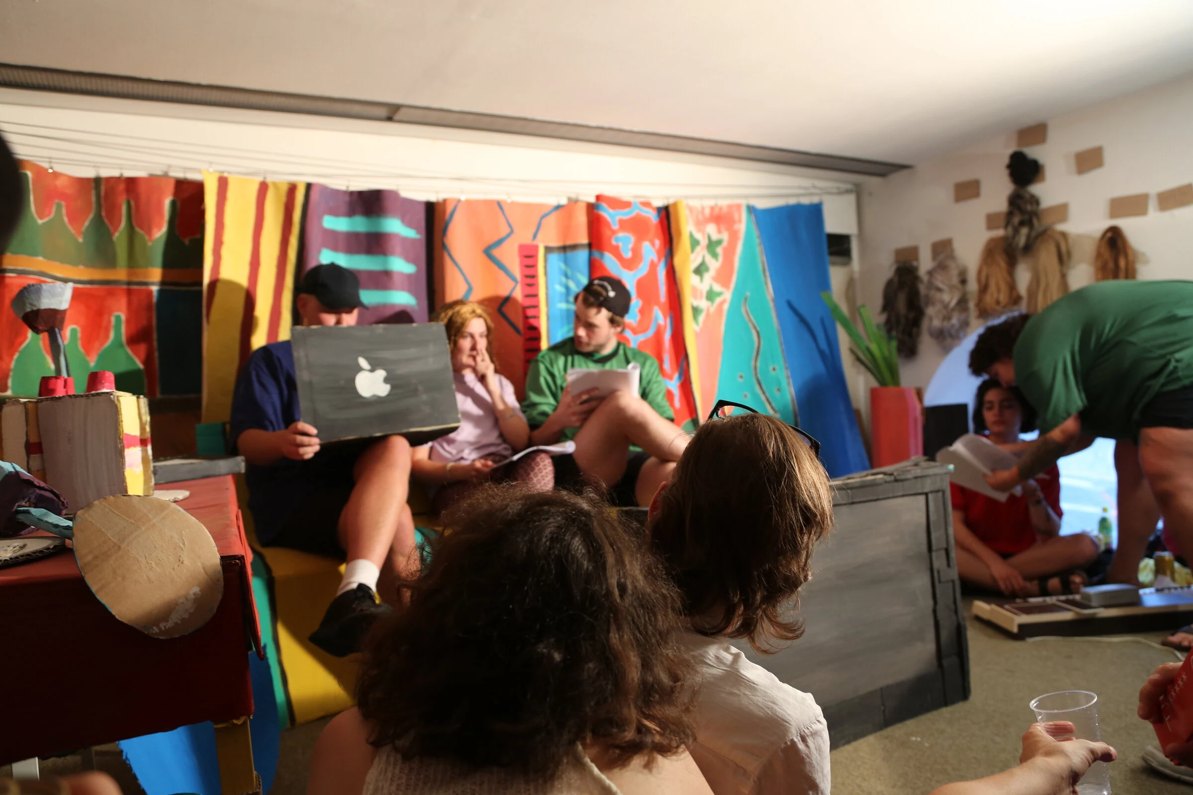A wide view of performers seated with scripts and a cardboard laptop, with audience members in the foreground.