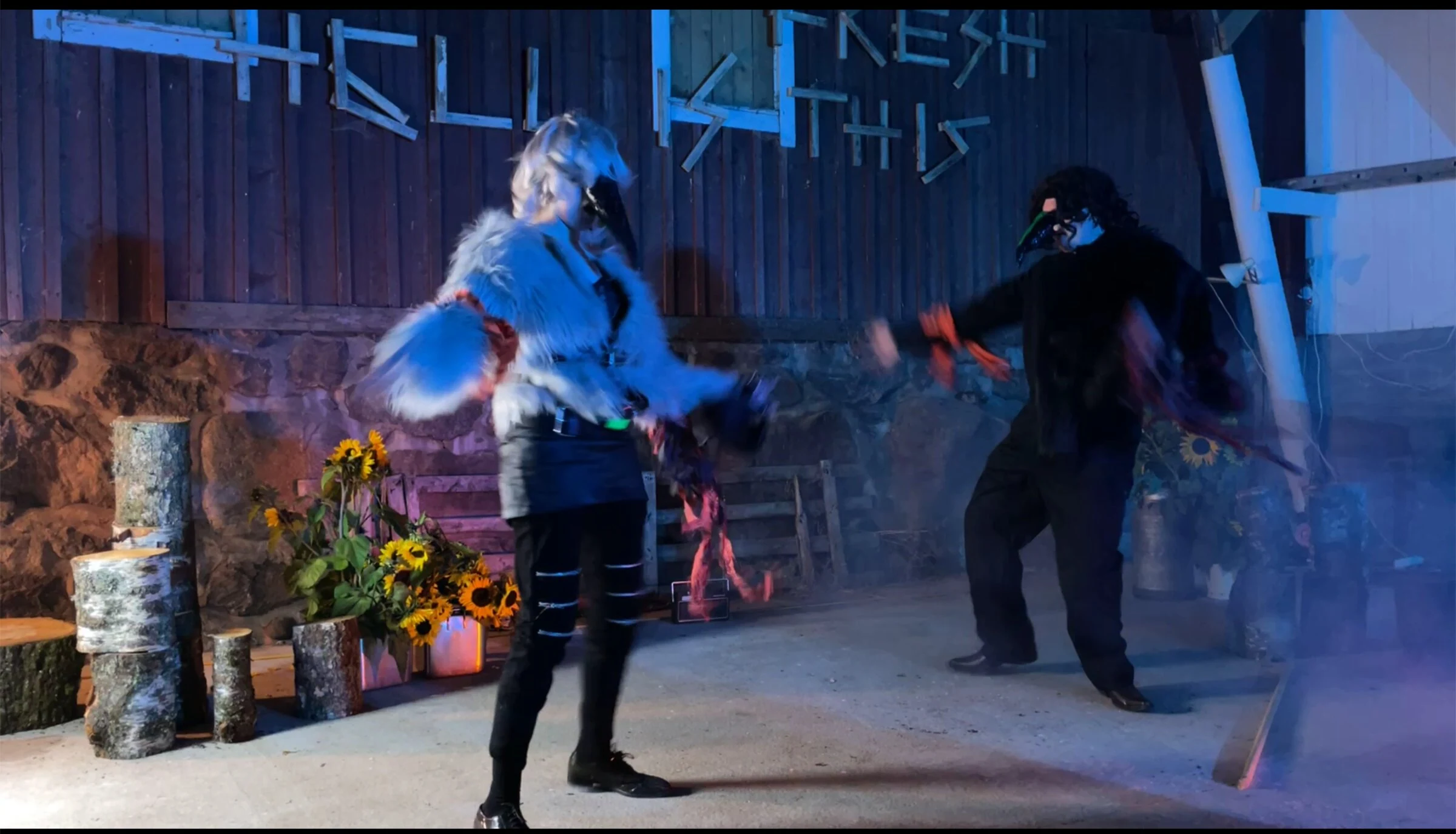 Two masked performers dancing beside sunflowers under blue lighting.