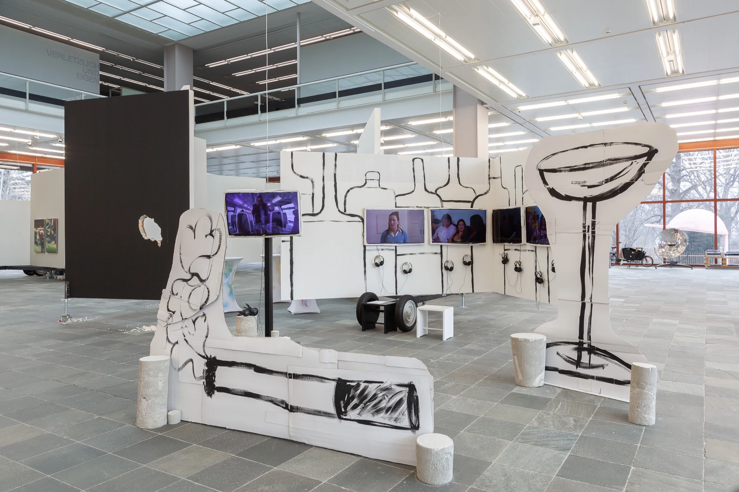 A wide view of LOCATION with video screens, bottle drawings and oversized cardboard sculptures.