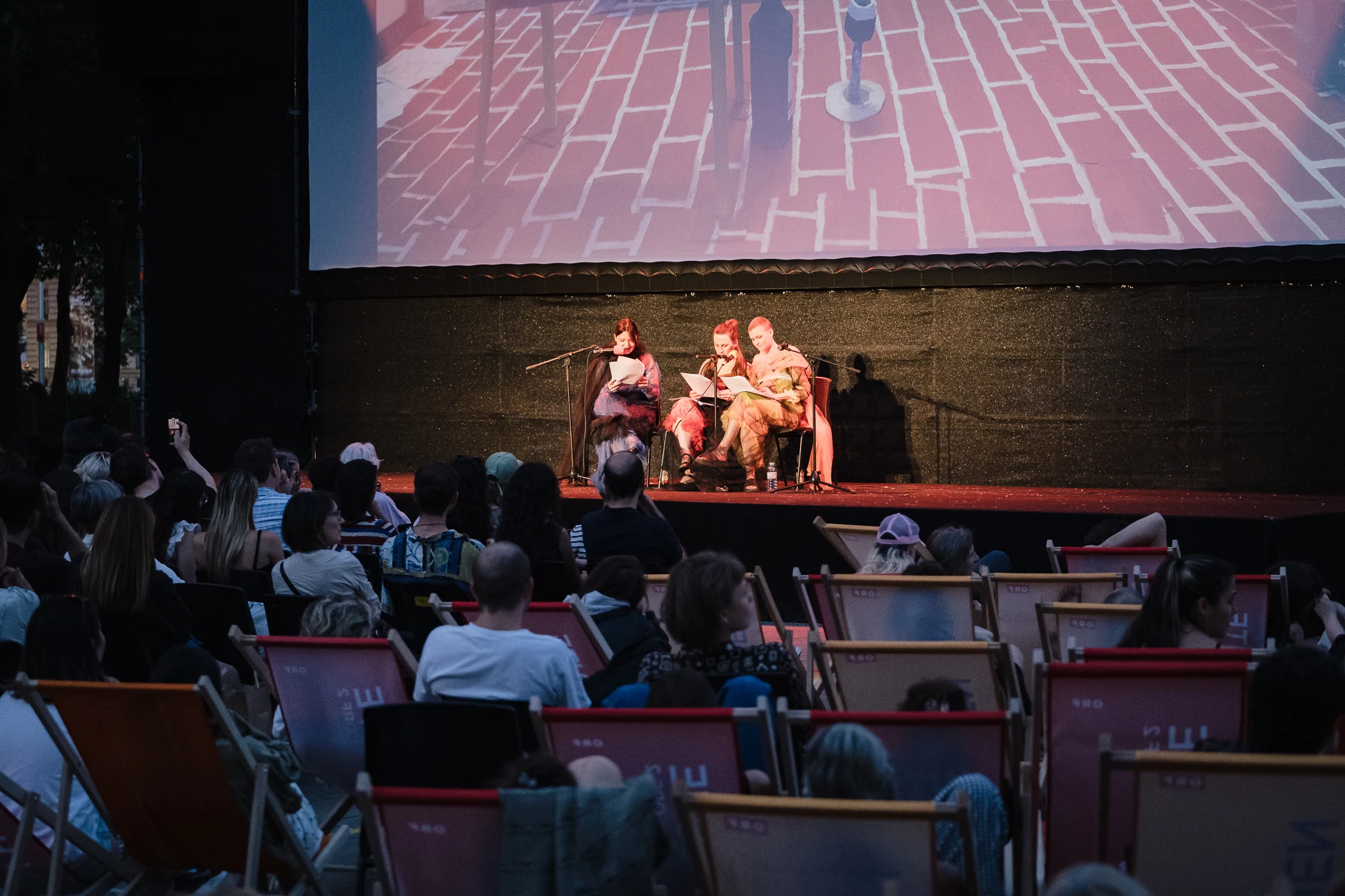 An outdoor audience watching three performers beneath a large projection screen.