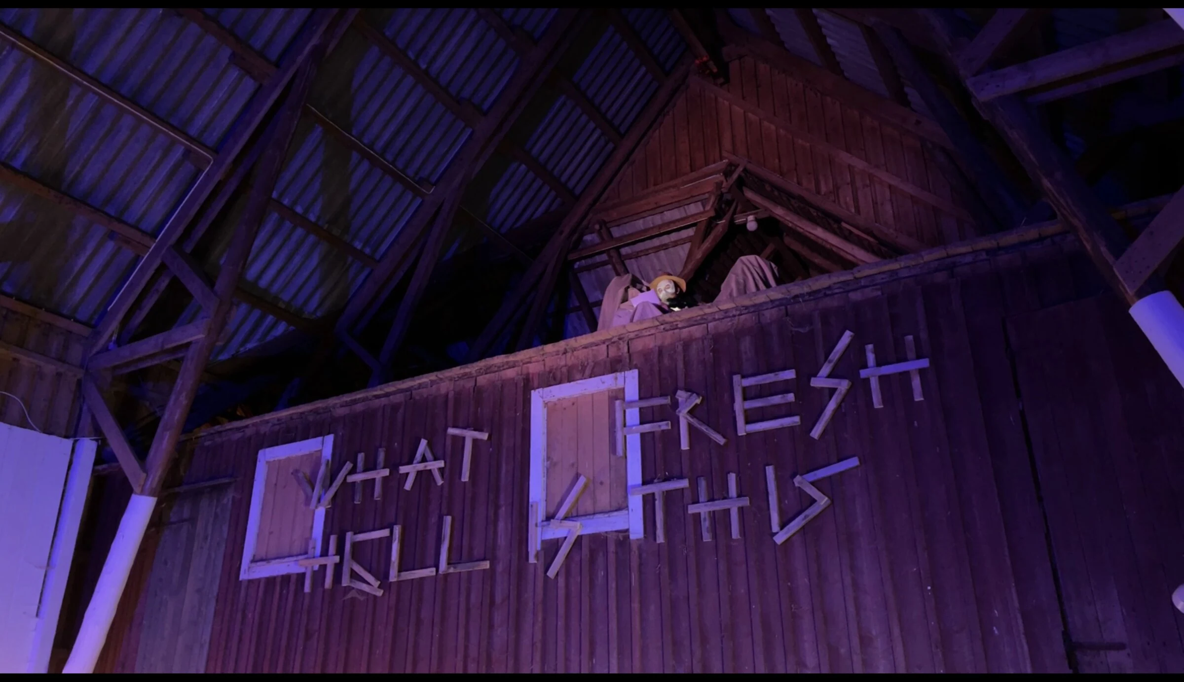 A masked performer reading from a loft above wooden lettering on the barn wall.