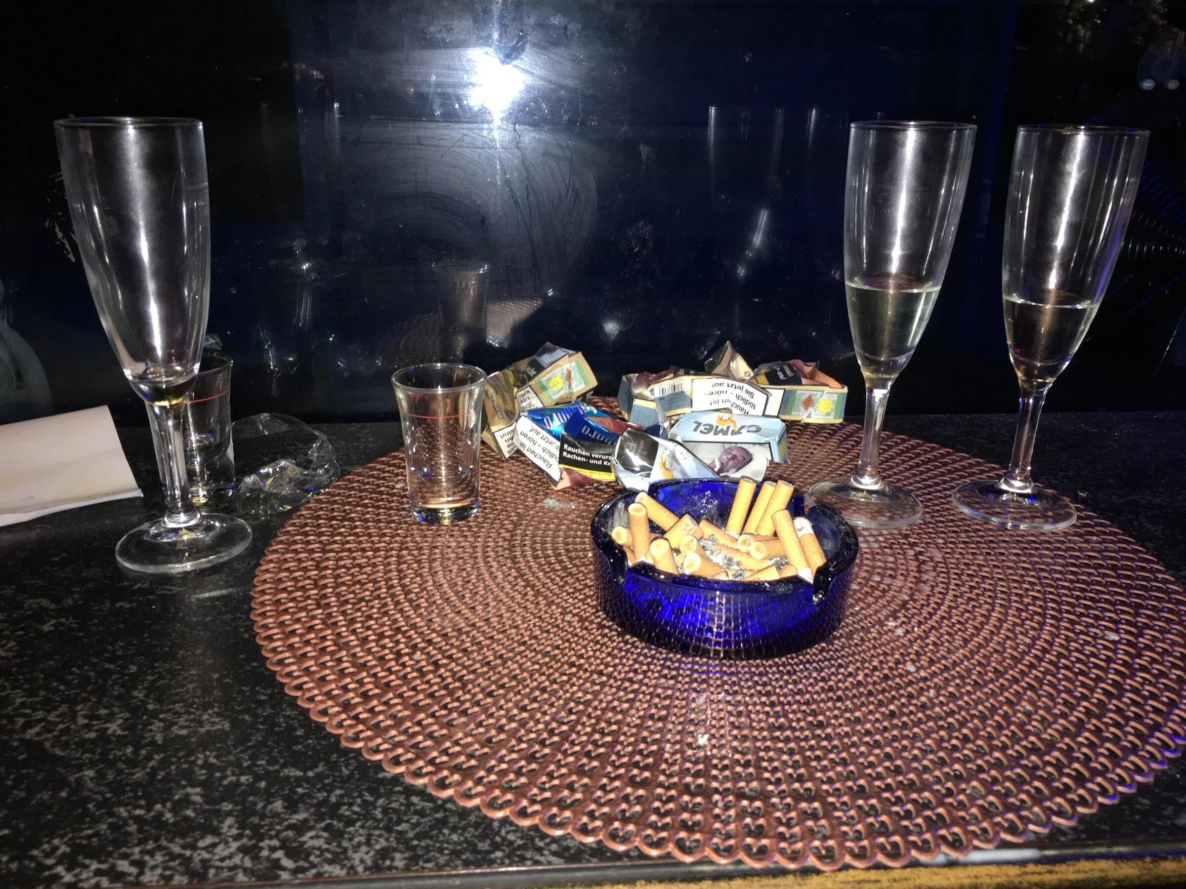 Drinking glasses, cigarette packets and a blue ashtray on a bar table.