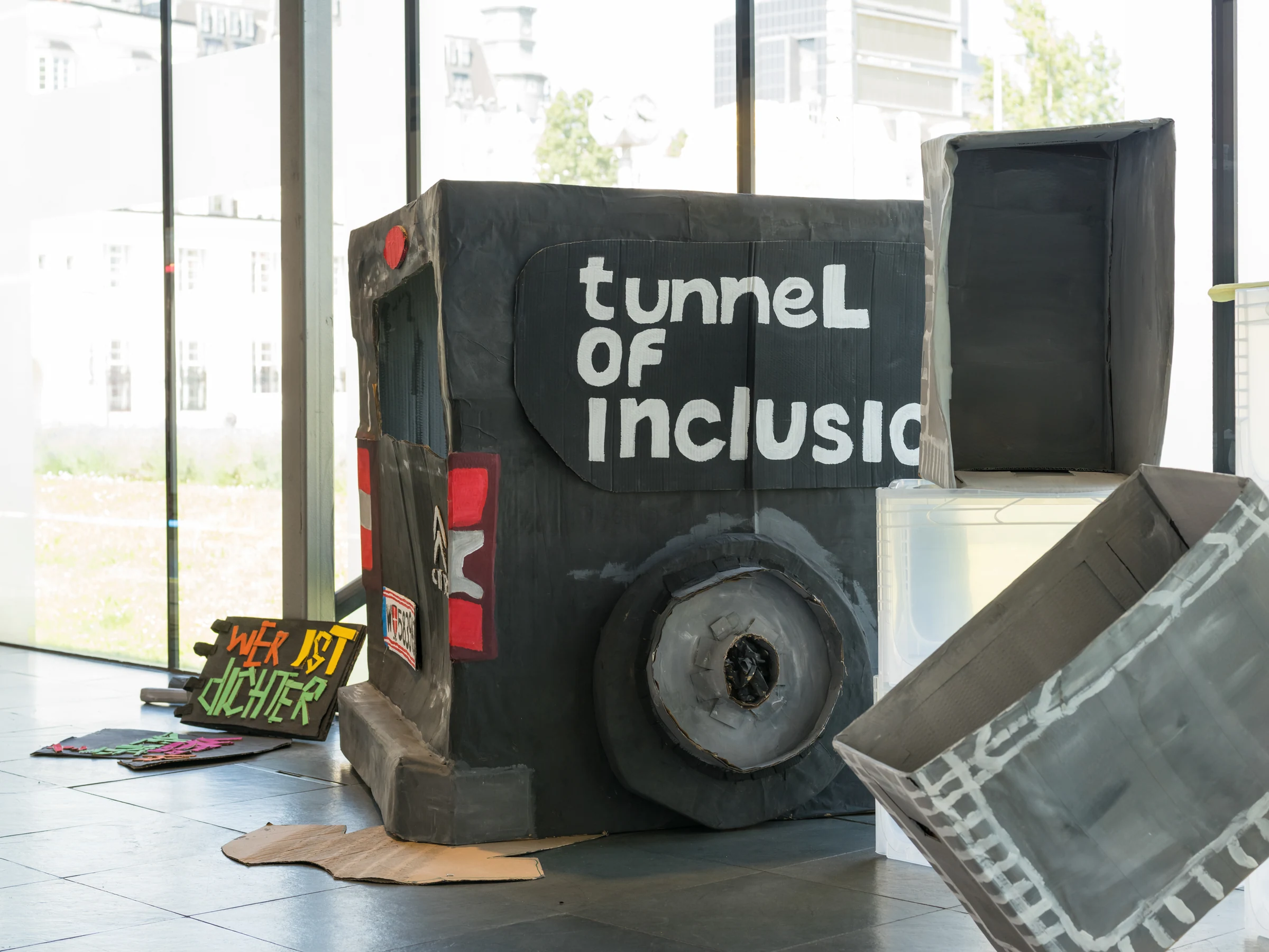 A grey cardboard installation with a wheel and a hand-painted Tunnel of Inclusion sign in a bright gallery.