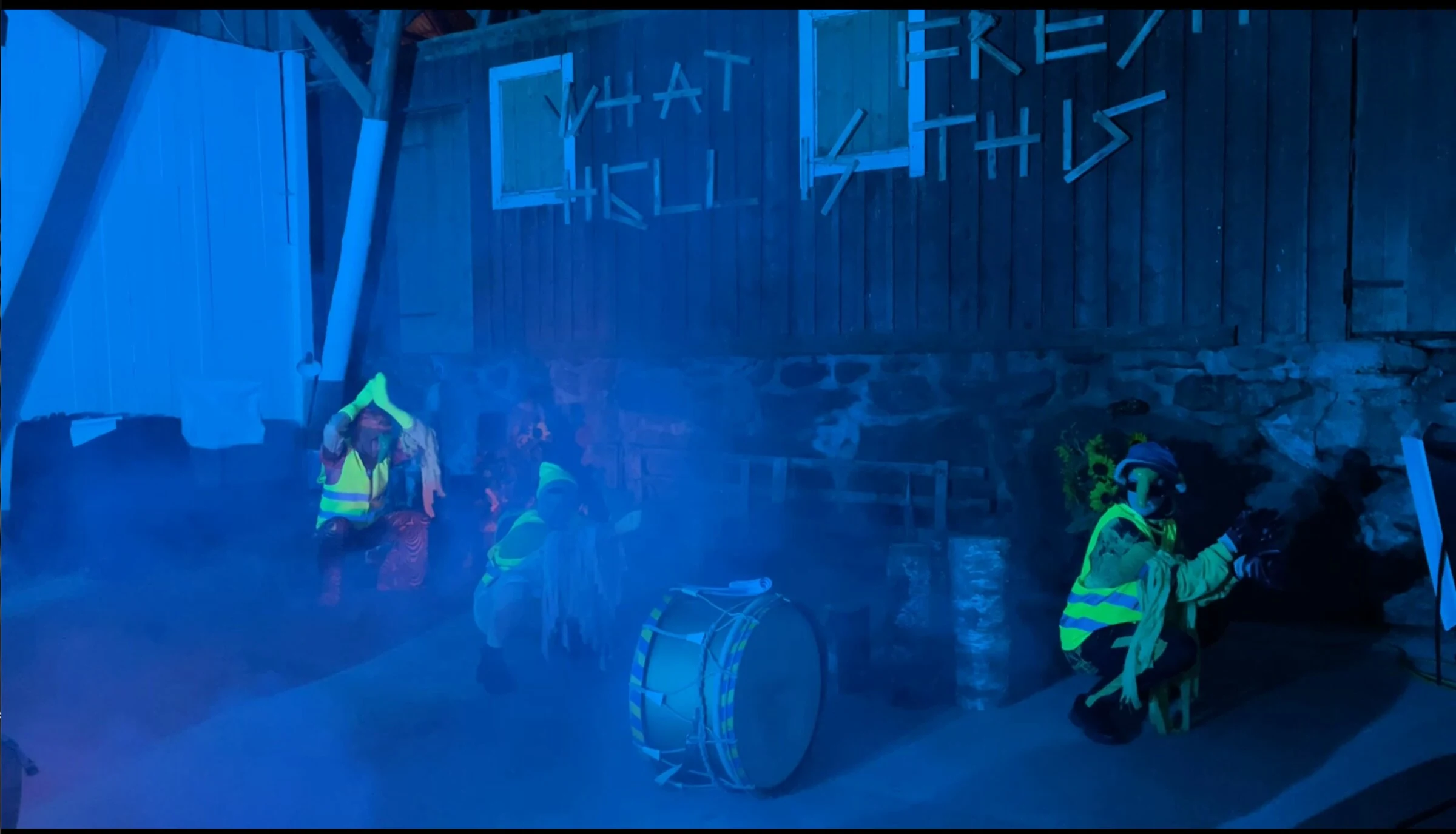 Three performers in fluorescent vests dancing in blue stage lighting and haze.