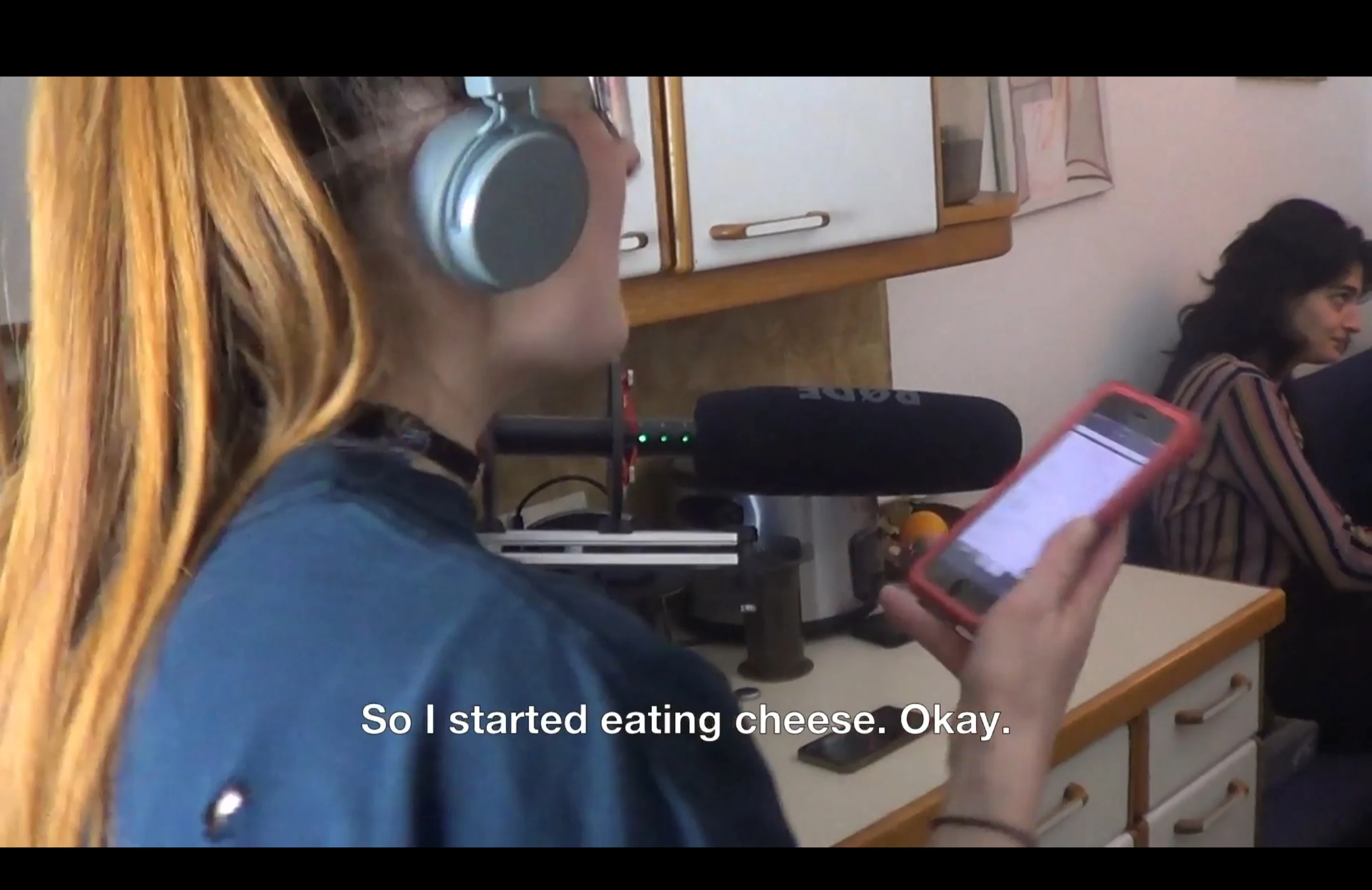 A crew member wearing headphones reads from a phone beside a microphone in the kitchen.