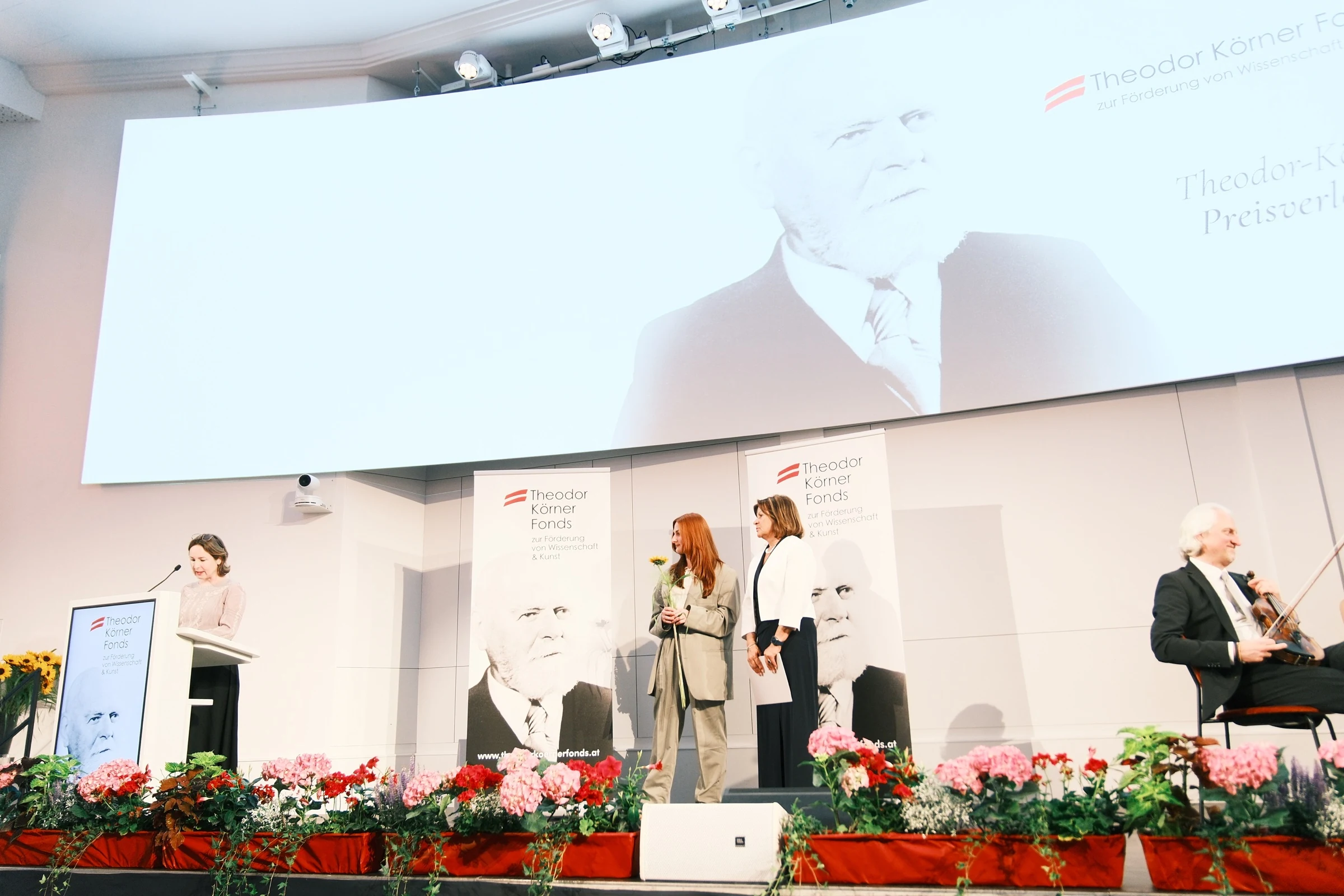 Charlotte Gash on stage during the Theodor Körner Prize ceremony, with a speaker and violinist.