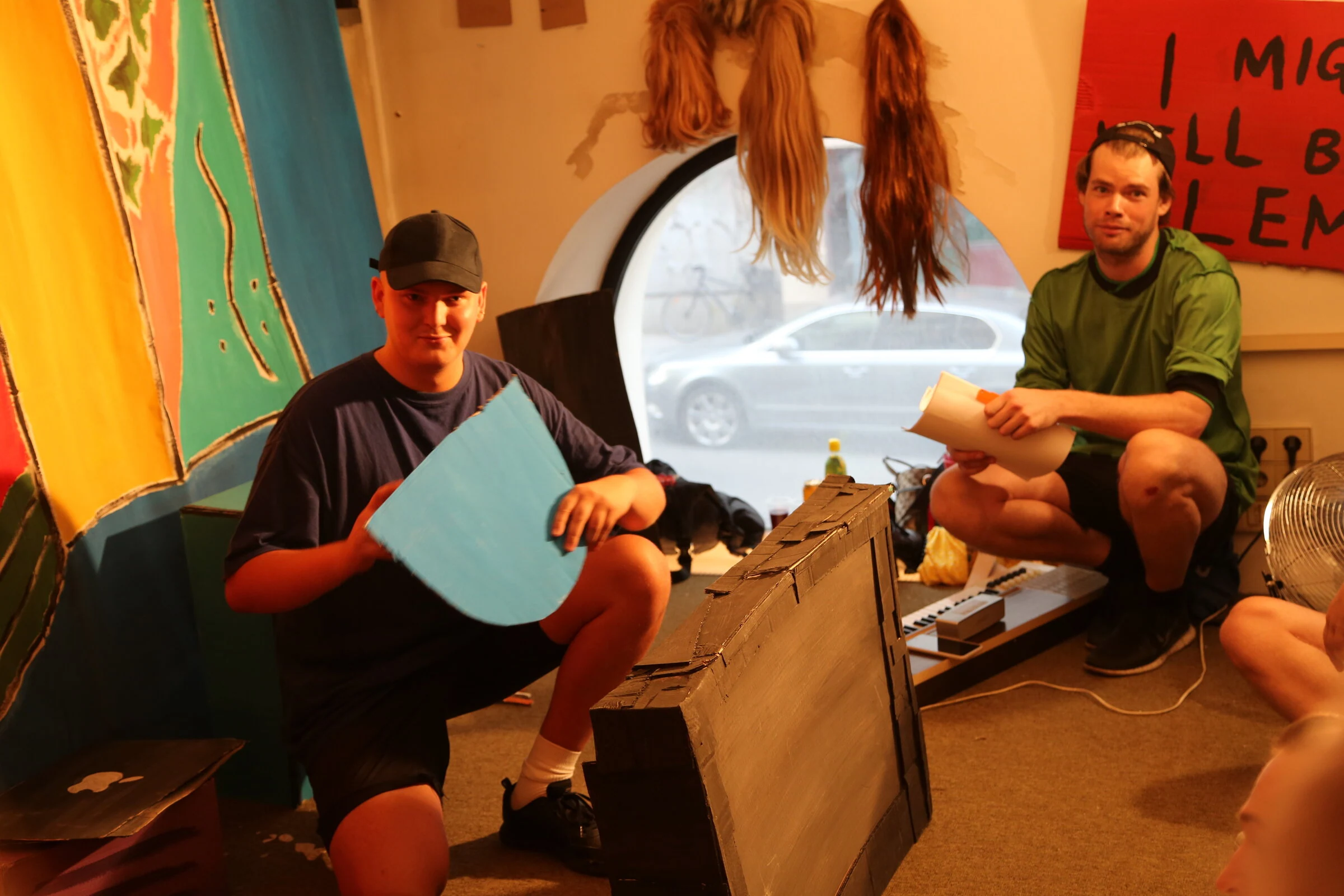 Two performers in caps holding a blue cardboard prop and a script beside a cardboard television and keyboard.