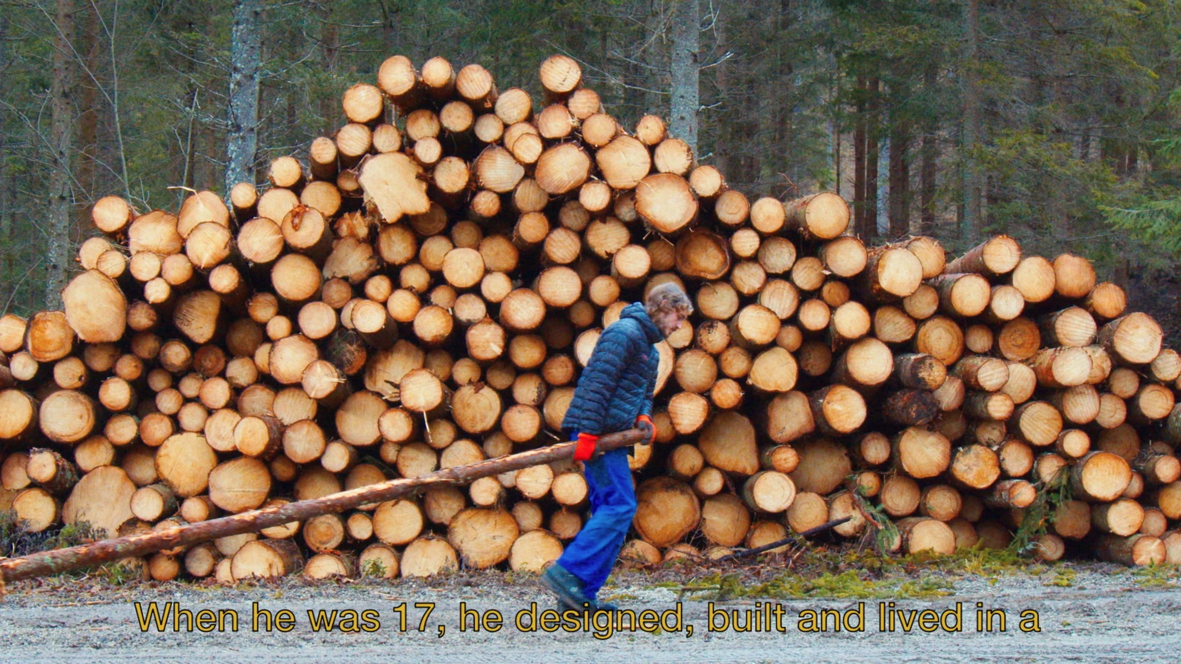A performer dragging a log past a large stack of timber.