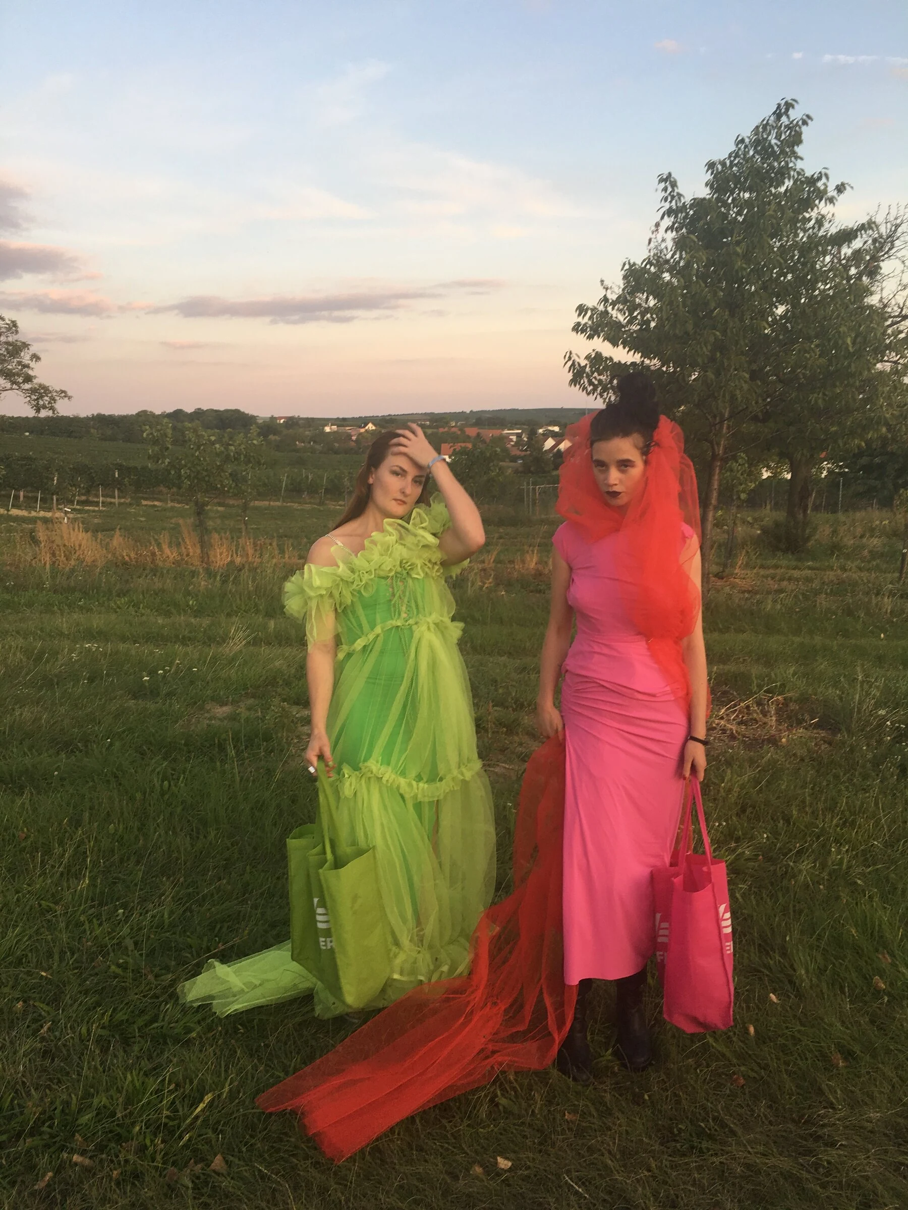 Two performers wearing green and pink dresses with long tulle trains in a field at sunset.