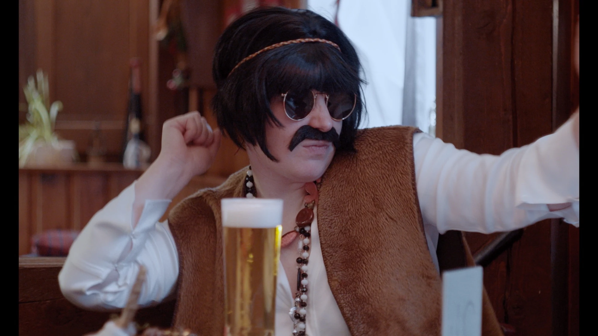 A character in a dark wig, sunglasses and false moustache sitting beside a glass of beer.