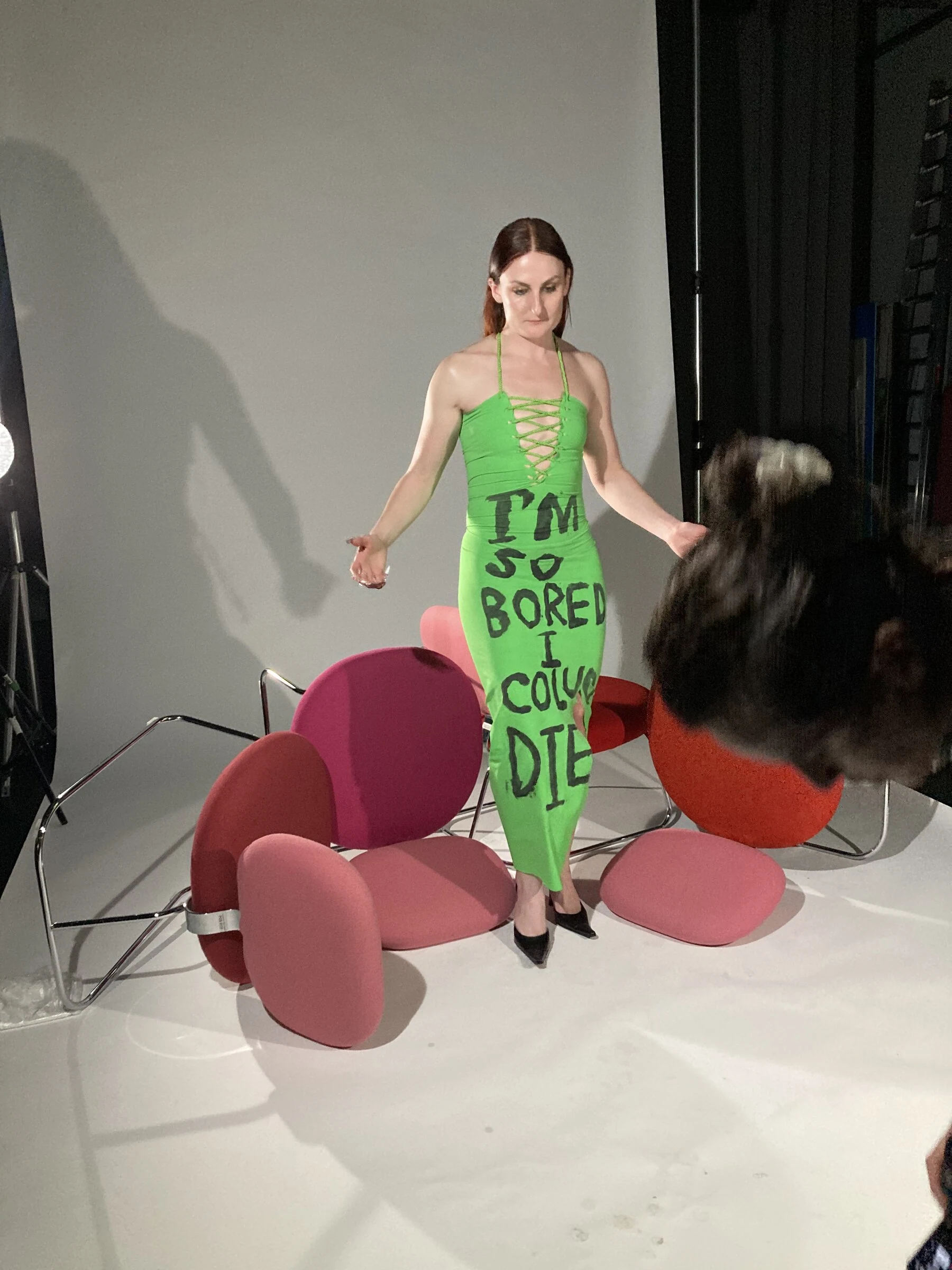 A studio photograph of Charlotte in a green dress with black lettering beside pink sculptural props.