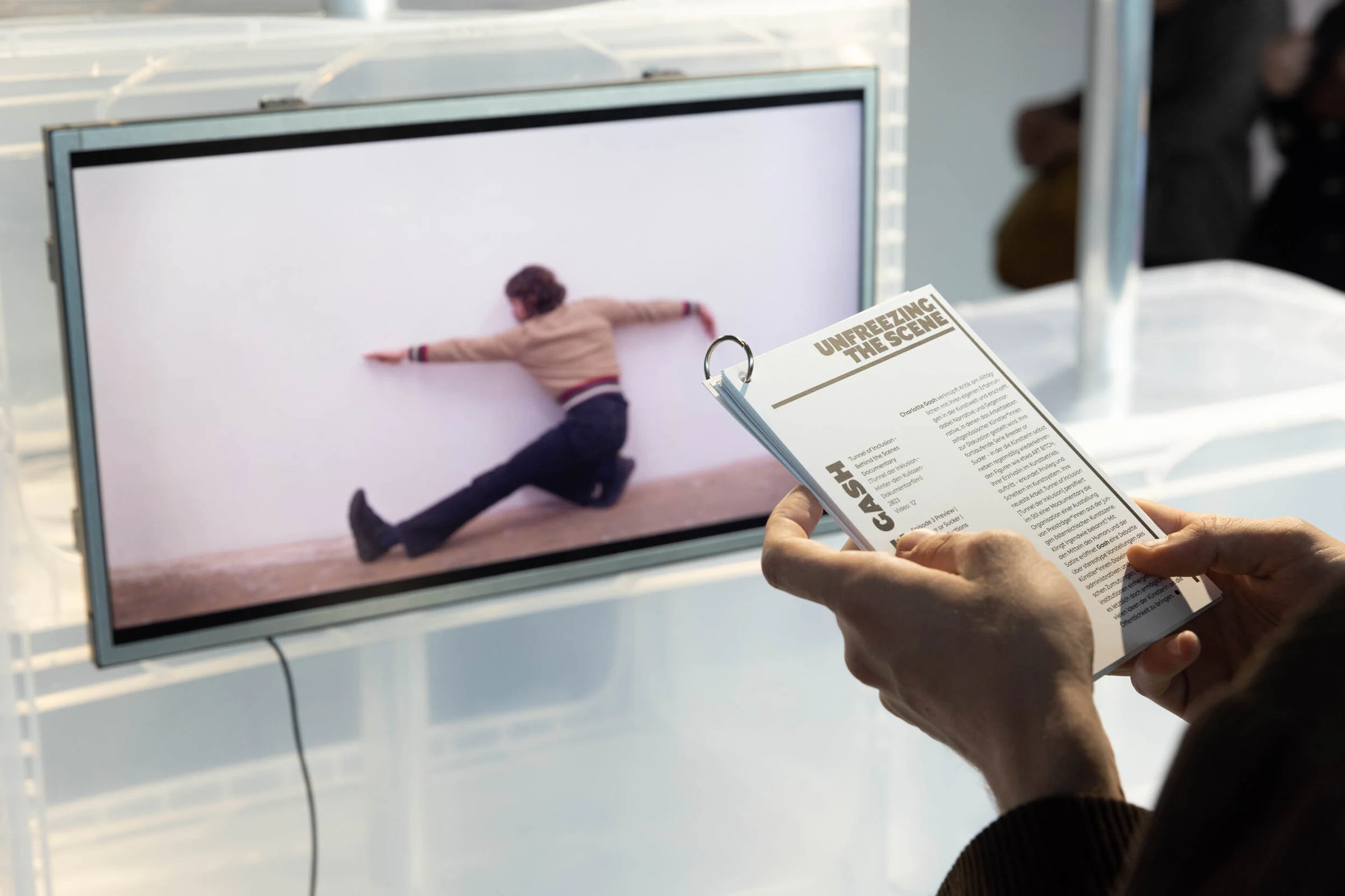 A visitor holding an Unfreezing the Scene exhibition booklet beside a screen showing a performer against a white wall.