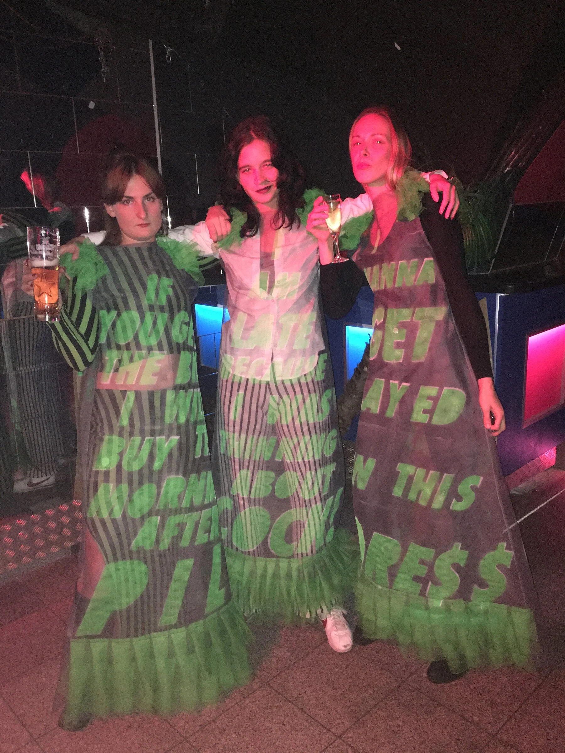 Three performers wearing dresses with projected green lettering under pink club lighting.