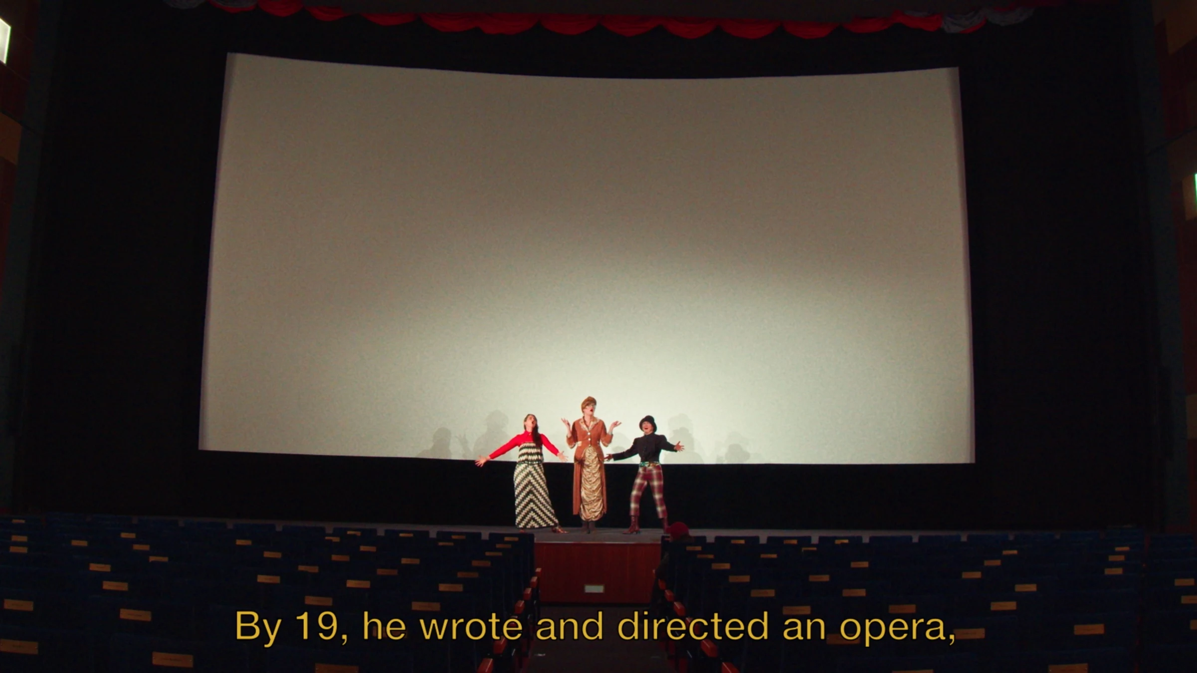 Three performers on a cinema stage beneath a large screen.