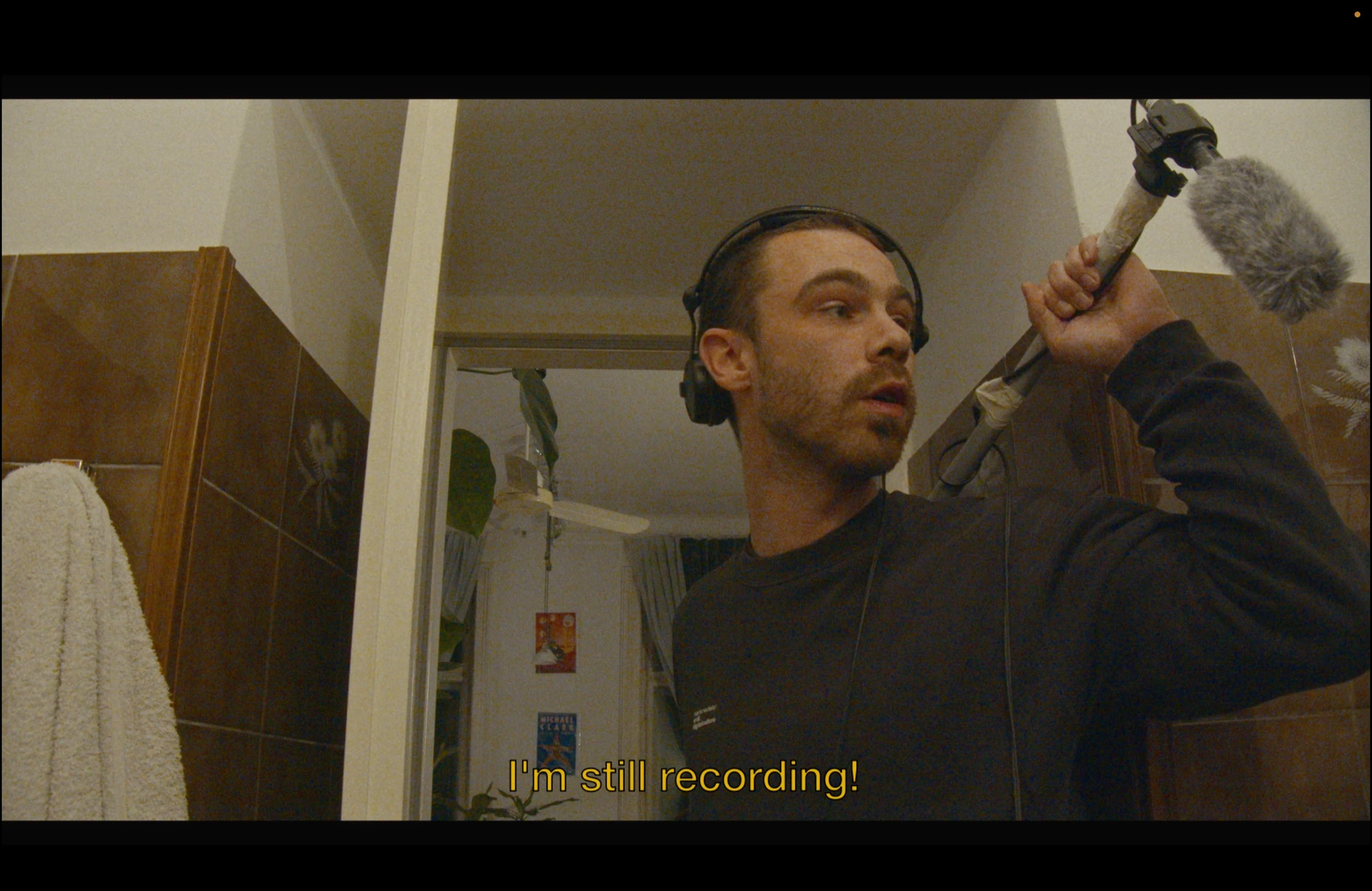 A sound recordist wearing headphones and holding a boom microphone in a tiled bathroom, subtitled I'm still recording!
