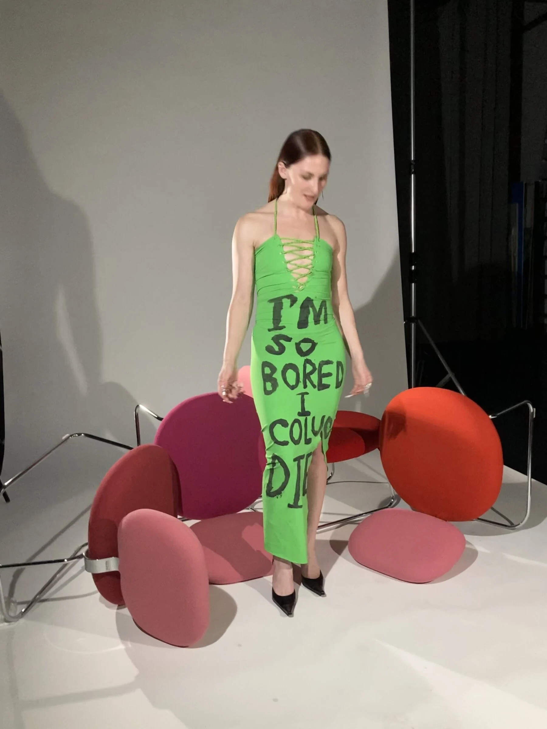 A performer wearing a green dress with black lettering in a studio with colourful sculptural props.